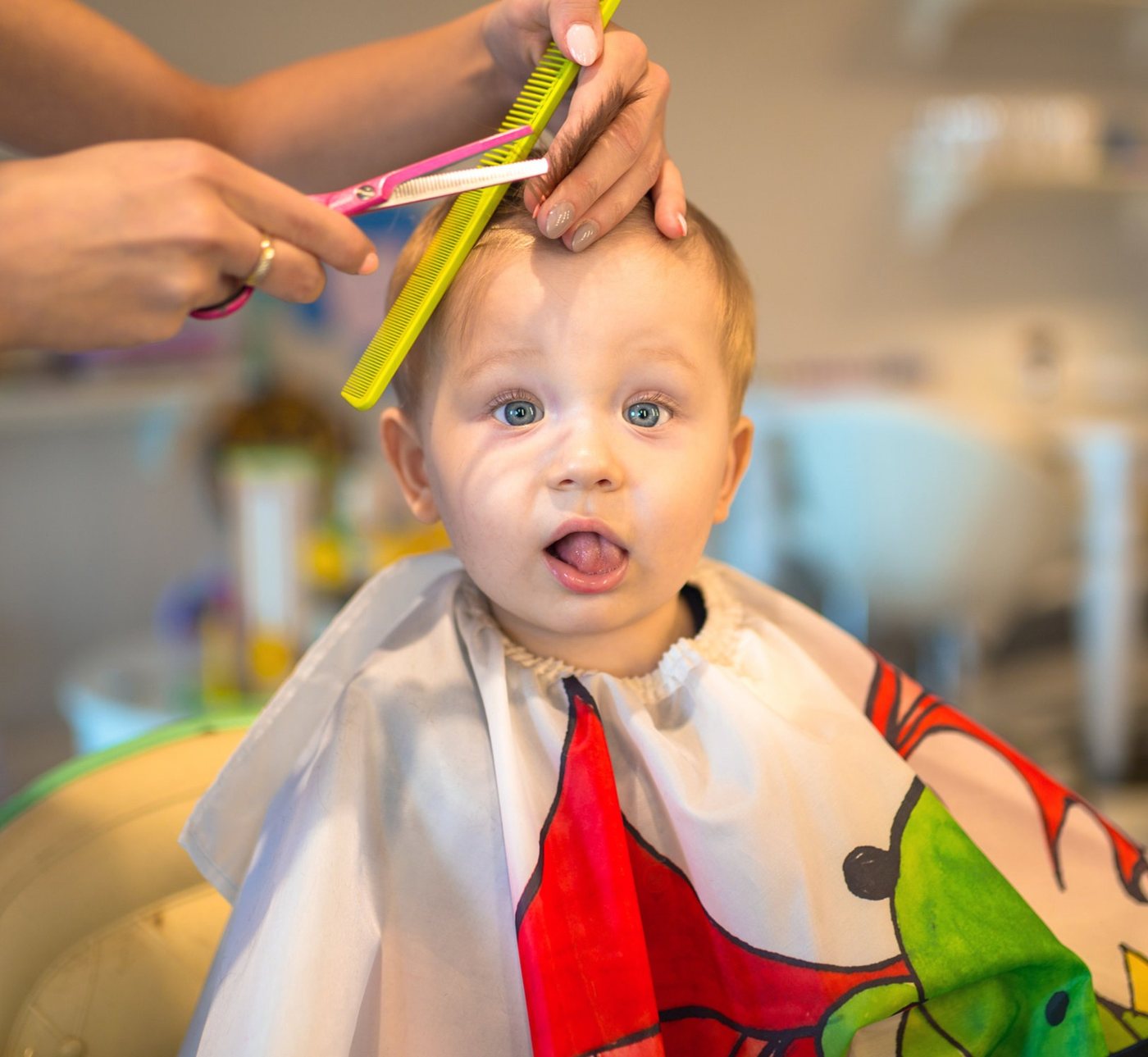 A toddler in a cape pulling a face while a stylist lifts a section of his hair with a comb and shears.