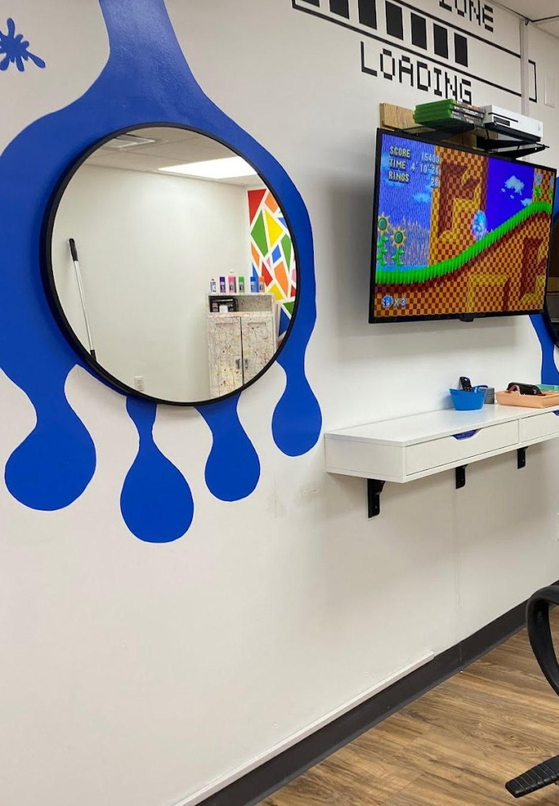 A round mirror mounted inside a large blue paint drip painted on the wall, next to a wall-mounted screen and game console.