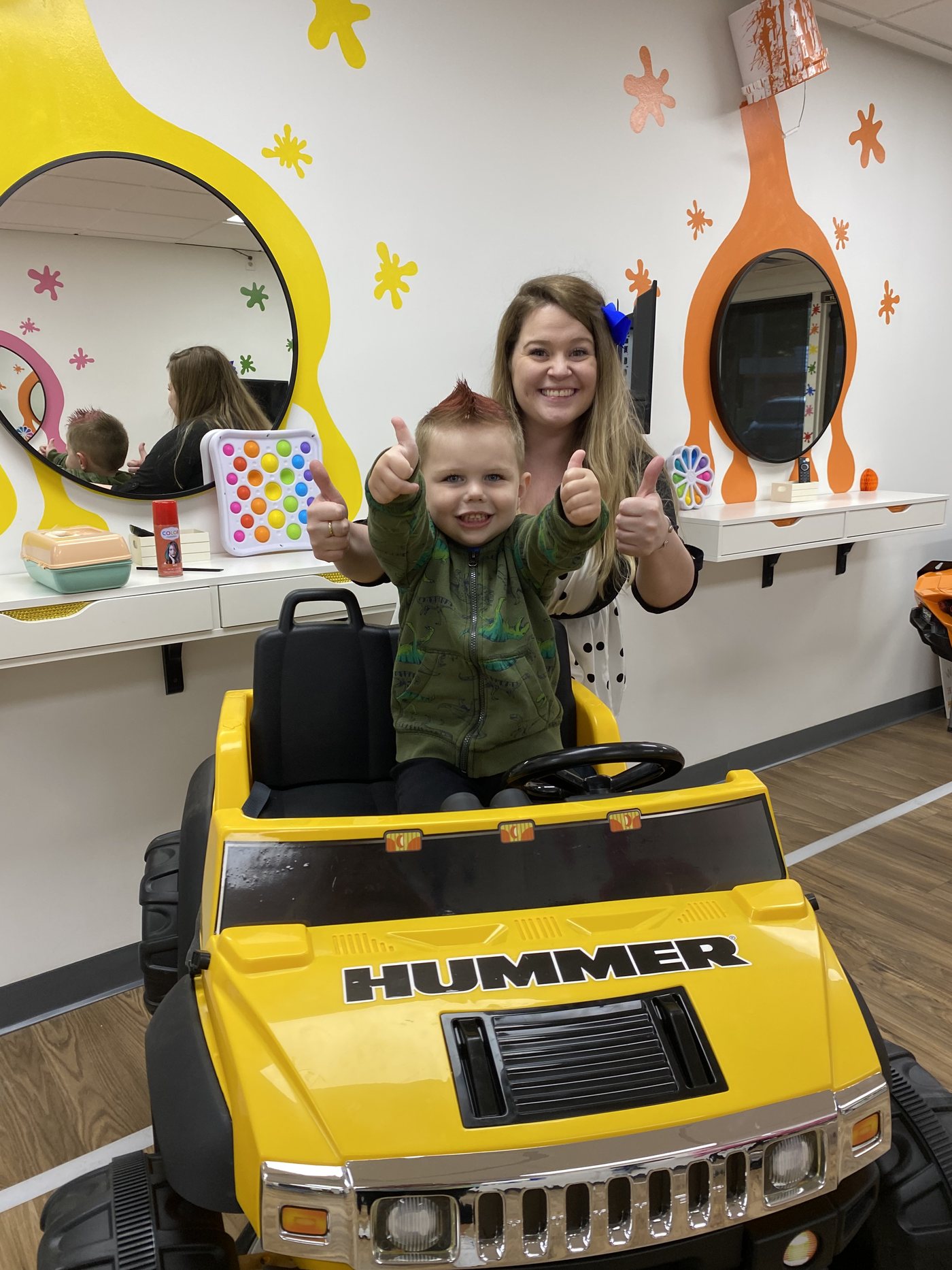 A yellow ride-on truck parked at a styling station, with round mirrors set inside orange and yellow painted splats on the wall behind it.