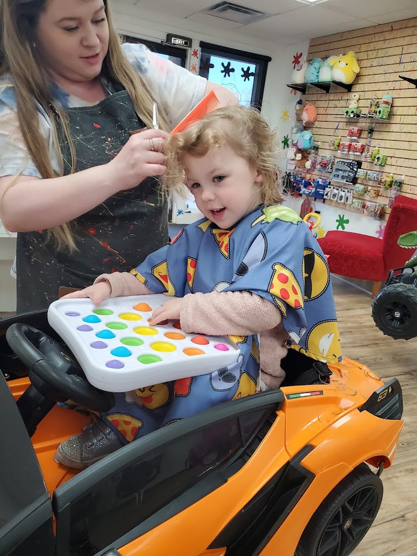 A toddler in an orange ride-on car holding a large pop-it fidget tray while a stylist trims her hair.